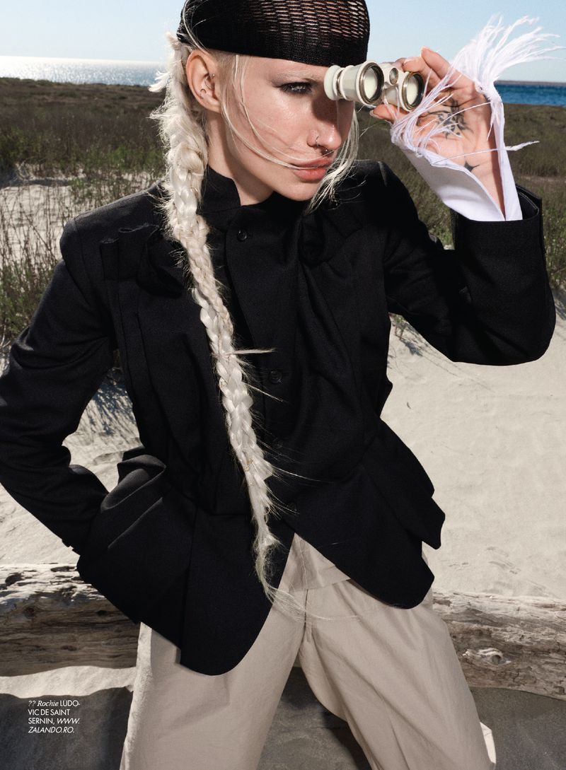 Fashion model with blonde braid and black headwear looking through binoculars outdoors in a black jacket and beige trousers.