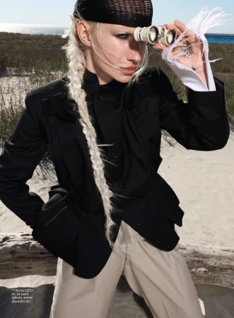 Fashion model with blonde braid and black headwear looking through binoculars outdoors in a black jacket and beige trousers.