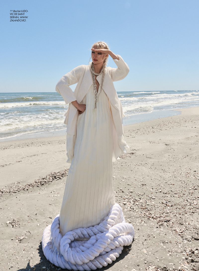 Woman wearing a long off-white pleated dress and jacket standing inside a coiled white rope on a sandy beach by the ocean.