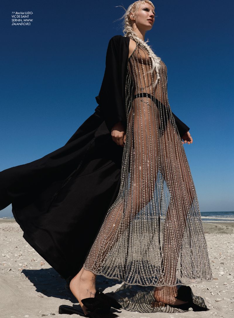 Model in sheer beaded dress and black coat walking on sandy beach against clear blue sky