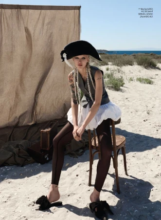 Editorial model wearing a black bicorne hat, grey vest, and white puff skirt seated on a wooden chair on a sandy beach.