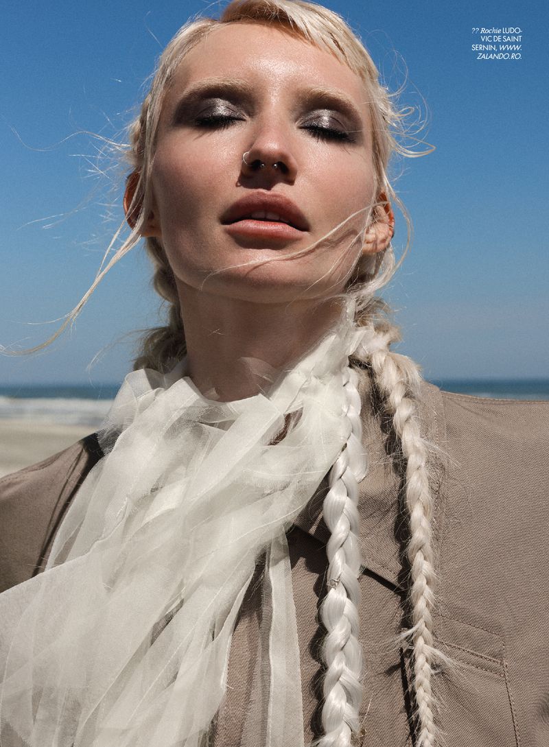 Close-up portrait of a woman with braided blonde hair wearing a white sheer scarf and taupe jacket against a beach and blue sky background.