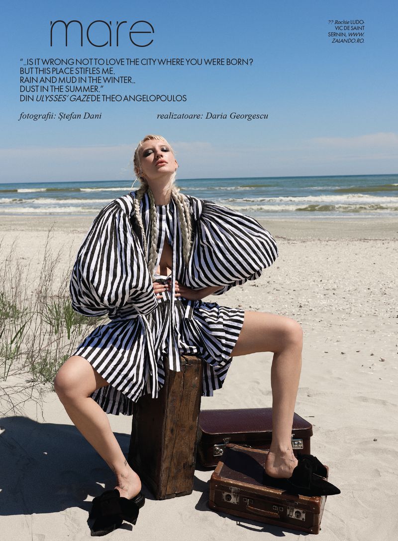 Model with braided blonde hair in a black and white striped dress sitting on a vintage suitcase on a sandy beach