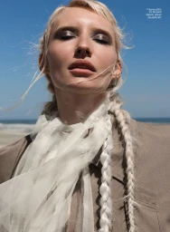 Close-up portrait of a woman with braided blonde hair wearing a white sheer scarf and taupe jacket against a beach and blue sky background.