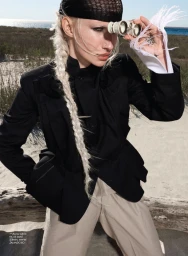 Fashion model with blonde braid and black headwear looking through binoculars outdoors in a black jacket and beige trousers.