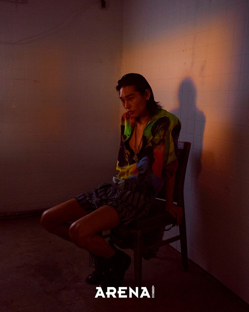 Model Taemin Park seated on a wooden chair in a printed shirt and striped shorts against a tiled wall in warm low-key lighting.