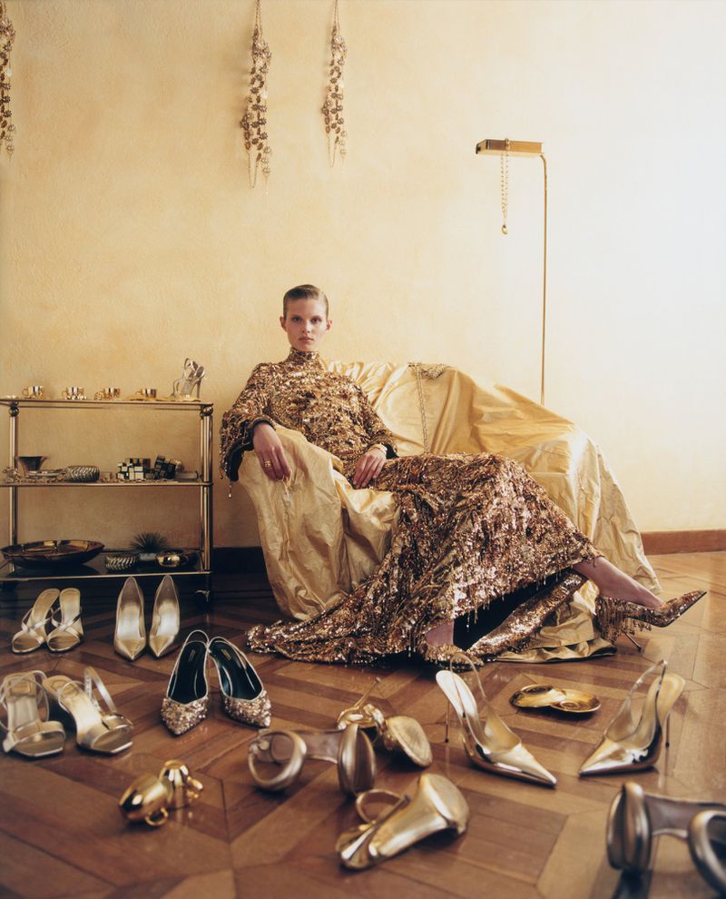 Model Ida Heiner in a shimmering gold gown seated on a draped chair surrounded by scattered metallic high heels on a wooden floor.