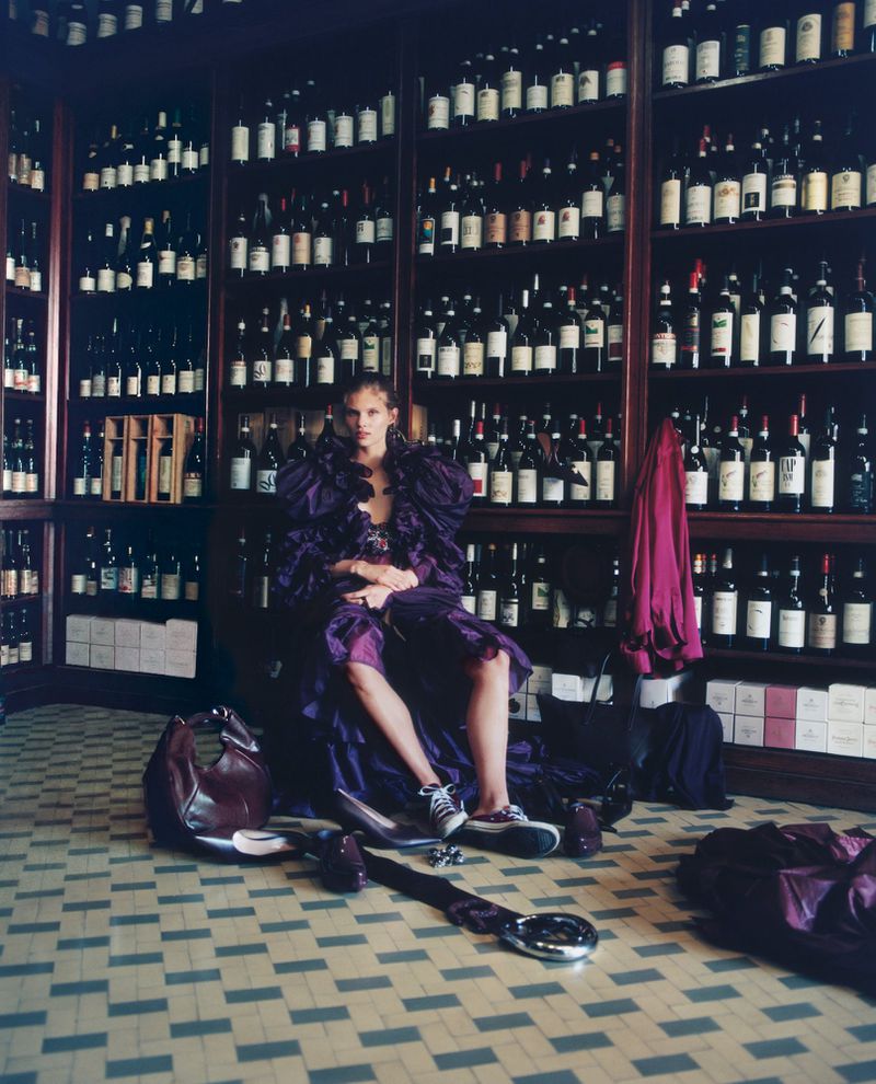 Model Ida Heiner in a voluminous purple dress seated inside a wine cellar with dark wood shelving lined with wine bottles.