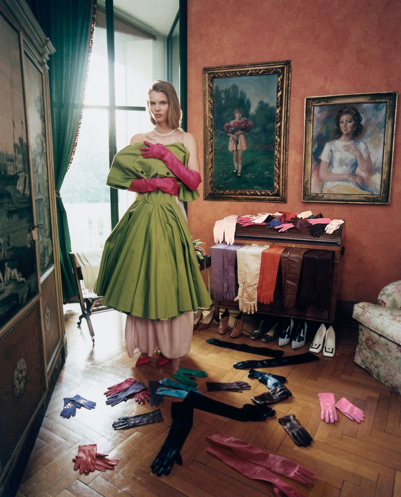 Model Ida Heiner wearing a voluminous olive green dress and pink opera gloves standing in an ornate room with gloves on the floor.