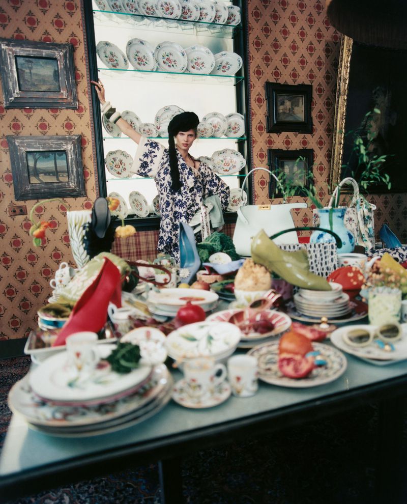 Model Ida Heiner in a patterned robe standing behind a lavish, cluttered table set with ceramic tableware, fruit, and high heels.