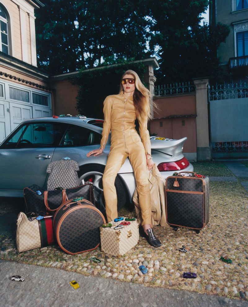 Model Ida Heiner in a camel leather outfit and visor sunglasses leaning against a silver sports car surrounded by luxury luggage.