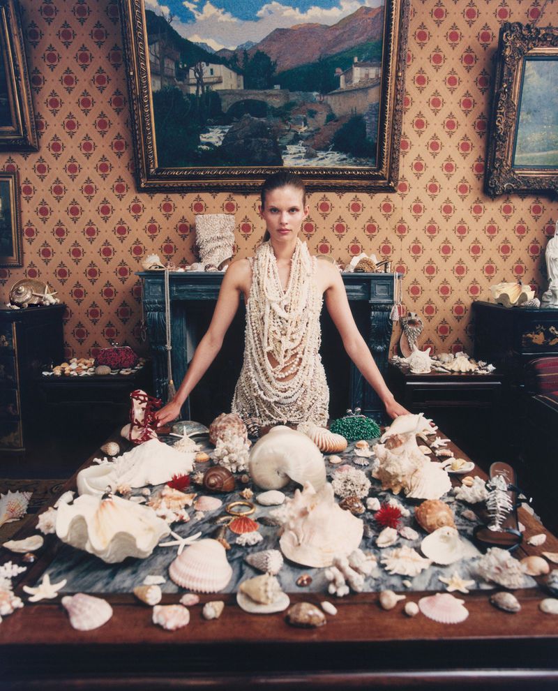 Model Ida Heiner wearing a layered white pearl net dress behind a table covered in sea shells and coral in a wallpapered room.