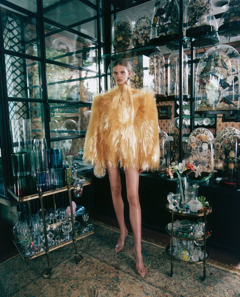 Model Ida Heiner in an exaggerated golden yellow feathered coat standing among glass cloches and display carts.