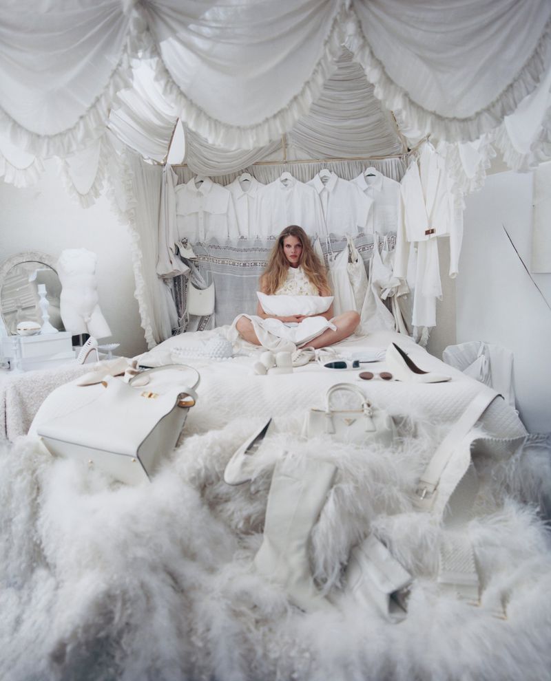 Model Ida Heiner sits cross-legged on an ornate white canopy bed surrounded by all-white clothing, handbags, and fluffy textures.
