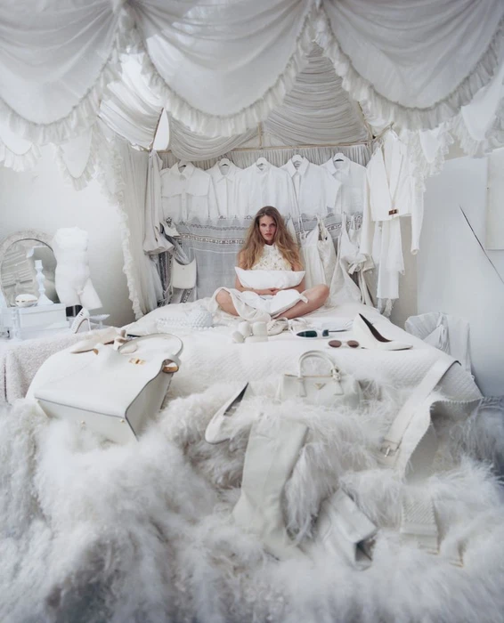 Model Ida Heiner sits cross-legged on an ornate white canopy bed surrounded by all-white clothing, handbags, and fluffy textures.
