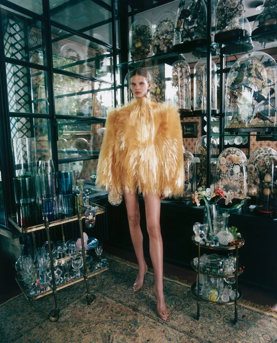 Model Ida Heiner in an exaggerated golden yellow feathered coat standing among glass cloches and display carts.