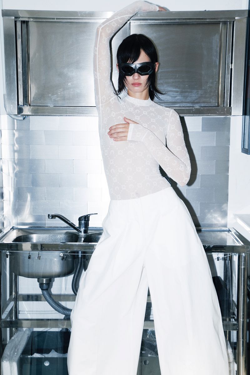 Model wearing dark wrap sunglasses and sheer patterned long-sleeve top standing in a commercial stainless steel kitchen