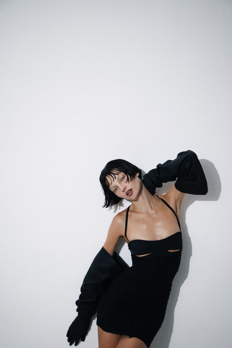 Model Zhenya Migovych wearing a black cut-out mini dress and long black gloves against a plain white studio background.