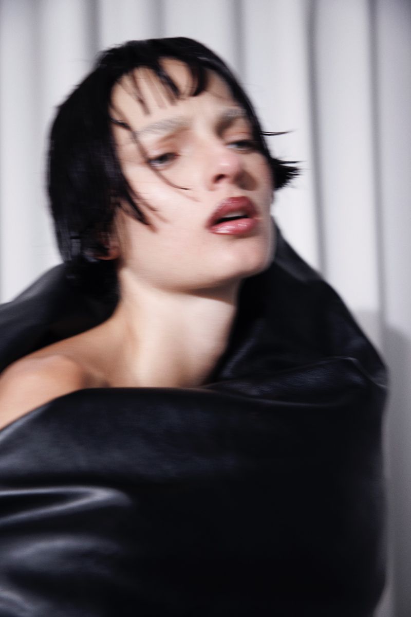 Model Zhenya Migovych wrapped in sculptural black fabric with dark fringe against a pleated backdrop.