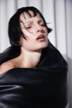 Model Zhenya Migovych wrapped in sculptural black fabric with dark fringe against a pleated backdrop.