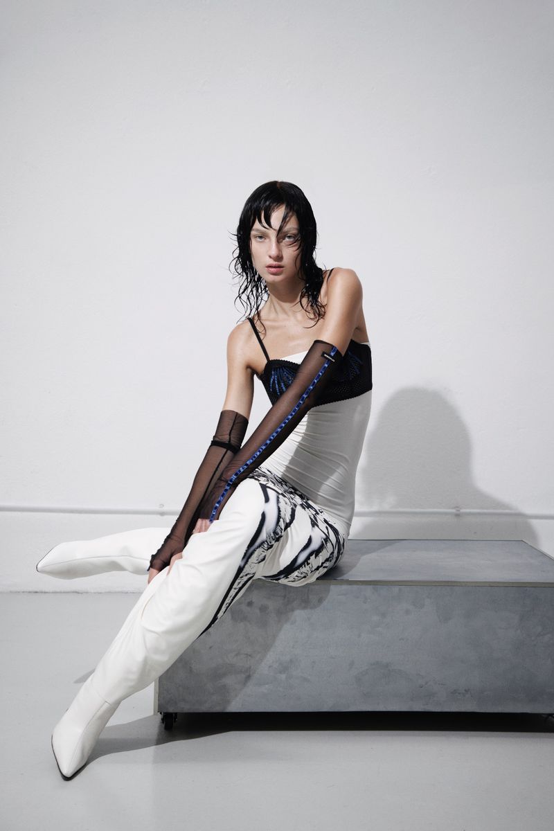 Model Zhenya Migovych seated on a grey bench in a white dress, black sheer gloves, and white boots against a white studio wall.