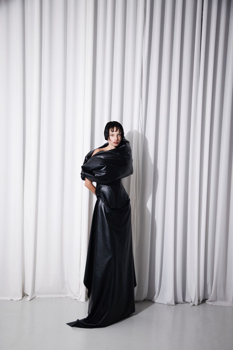Model Zhenya Migovych posing in a sculptural black leather gown in front of white pleated drapes.