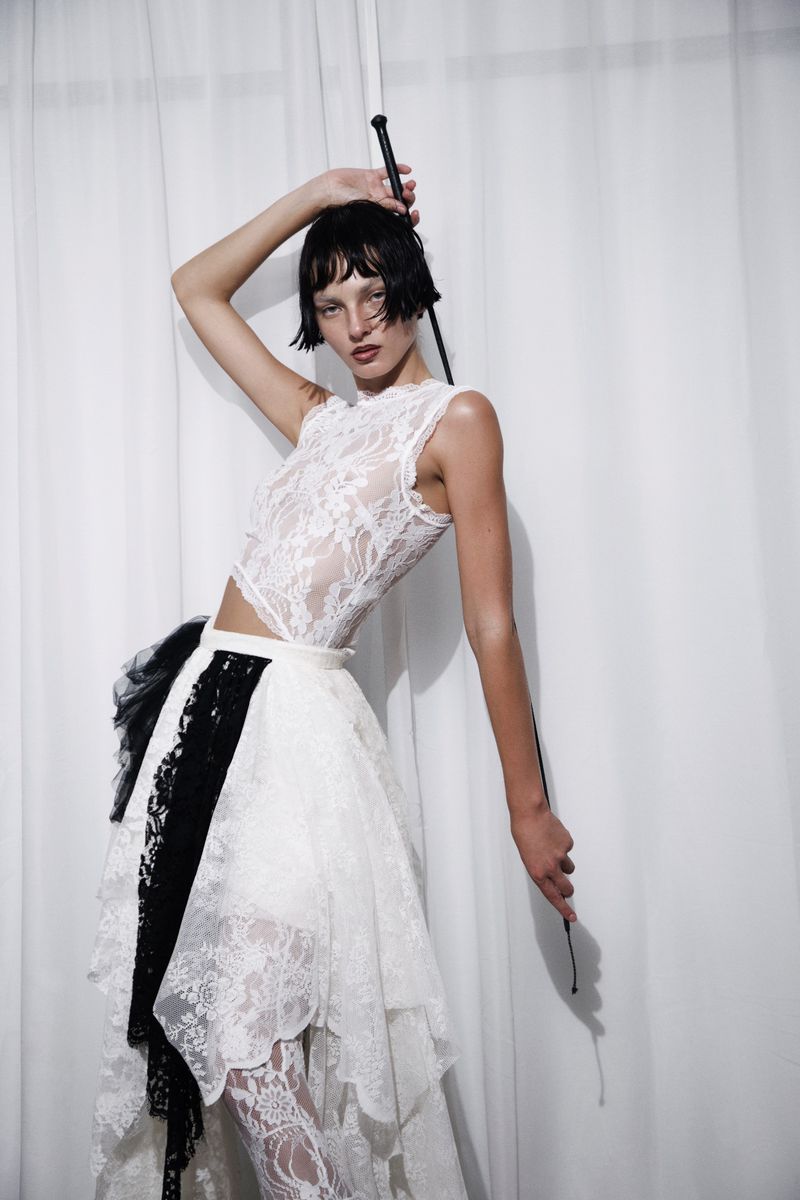 Model Zhenya Migovych posing in a white lace crop top and tiered skirt while holding a riding crop against white curtains.