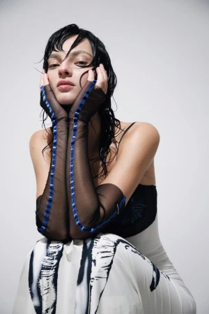 Model Zhenya Migovych posing with hands framing her face in sheer black gloves with blue trim against a plain white studio background.