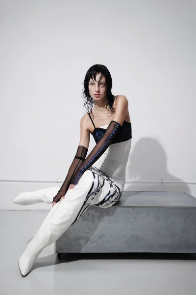 Model Zhenya Migovych seated on a grey bench in a white dress, black sheer gloves, and white boots against a white studio wall.
