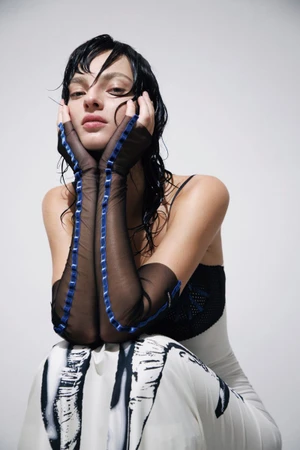 Model Zhenya Migovych posing with hands framing her face in sheer black gloves with blue trim against a plain white studio background.