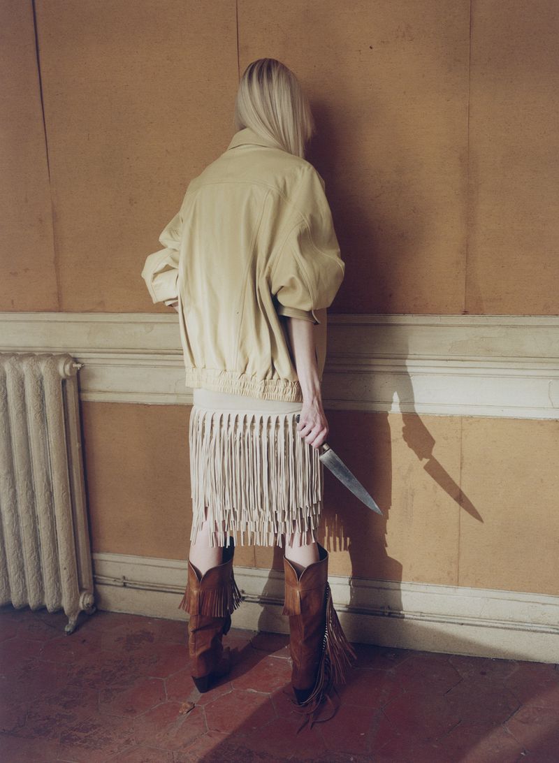 A woman seen from behind facing a wall, wearing a pale jacket, fringe skirt, and brown cowboy boots while holding a large knife.