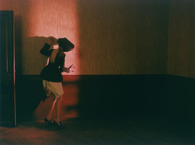 Model Malin Rudnick poses in a backless black jacket, cream skirt, and high heels against a red-lit textured wall.