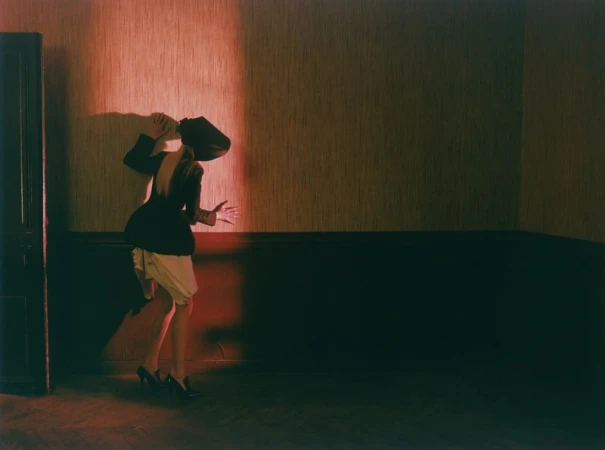Model Malin Rudnick poses in a backless black jacket, cream skirt, and high heels against a red-lit textured wall.
