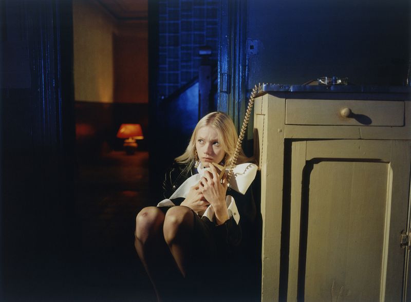 Model Malin Rudnick sits crouched on a dark floor clutching a corded telephone handset next to a wooden cabinet.
