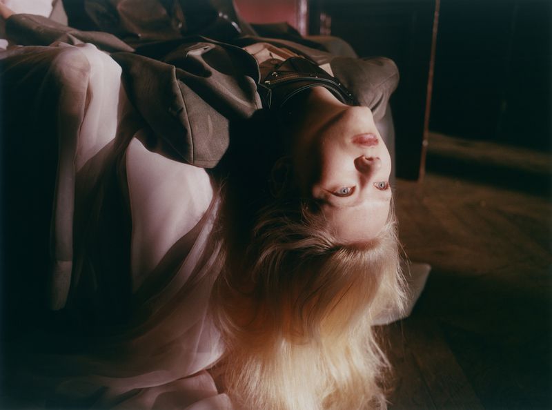 Model Malin Rudnick lying inverted with blonde hair draped downwards, wearing an oversized jacket and layered fabric.