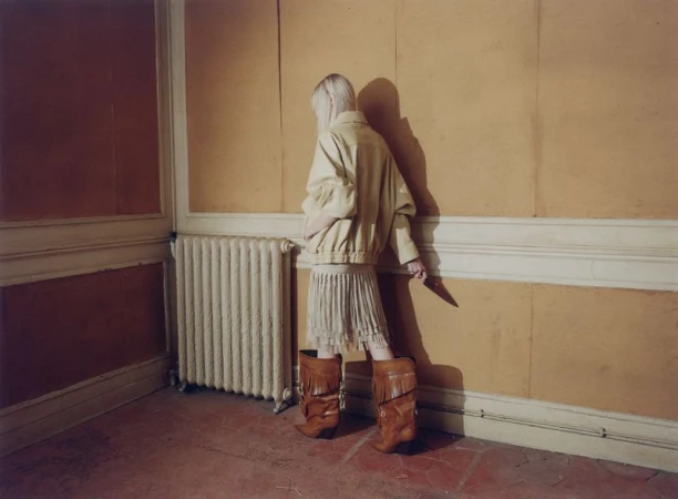 Model with blonde hair wearing fringed boots and an oversized jacket standing in a room with mustard walls.