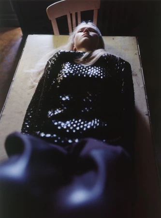 Model Malin Rudnick lying supine on a wooden table wearing a black sequined top, shot from a low foreshortened angle.