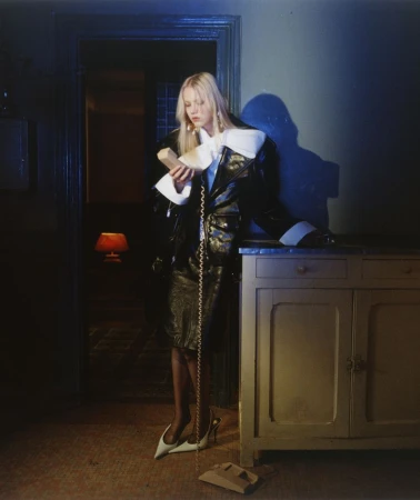 Model Malin Rudnick in a black patent coat with white collar accents holding a landline phone receiver beside a wooden cabinet.