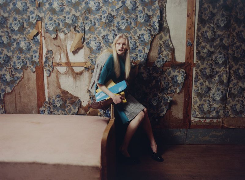 Model with long blonde hair holding books and bags seated in a derelict room with peeling floral wallpaper
