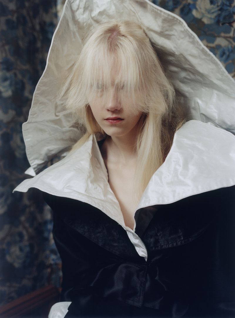 Model Malin Rudnick wearing an oversized sculptural white collar and black jacket against a patterned floral backdrop.