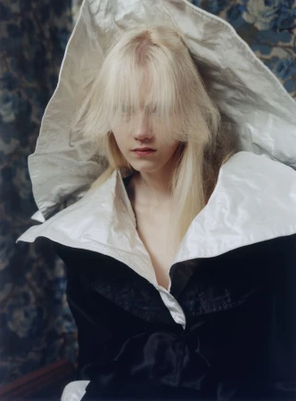 Model Malin Rudnick wearing an oversized sculptural white collar and black jacket against a patterned floral backdrop.
