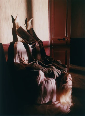 Model Malin Rudnick lying upside down on an armchair draped in fabric with legs resting against a wall next to a wooden door.