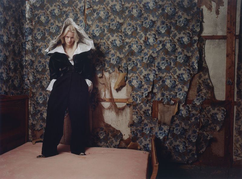Model Malin Rudnick wearing an oversized black outfit with white collar and hood, standing on a bed against torn floral wallpaper.