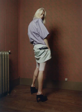 Model Malin Rudnick poses looking over her shoulder wearing a lilac short-sleeve shirt and draped skirt in a room with wallpaper.