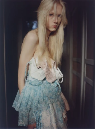 Blonde model Malin Rudnick in a tiered light blue lace and pastel dress standing in a softly lit interior doorway.