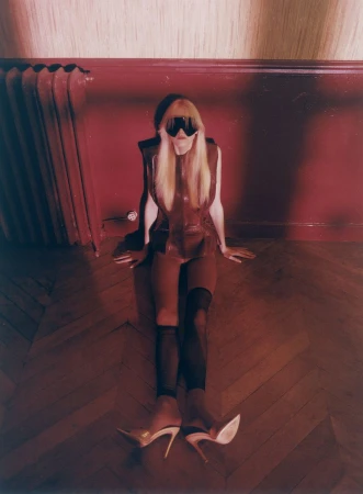 Model Malin Rudnick seated on a parquet wooden floor wearing a brown top, black visor sunglasses, and pointed high heels against a red wall.