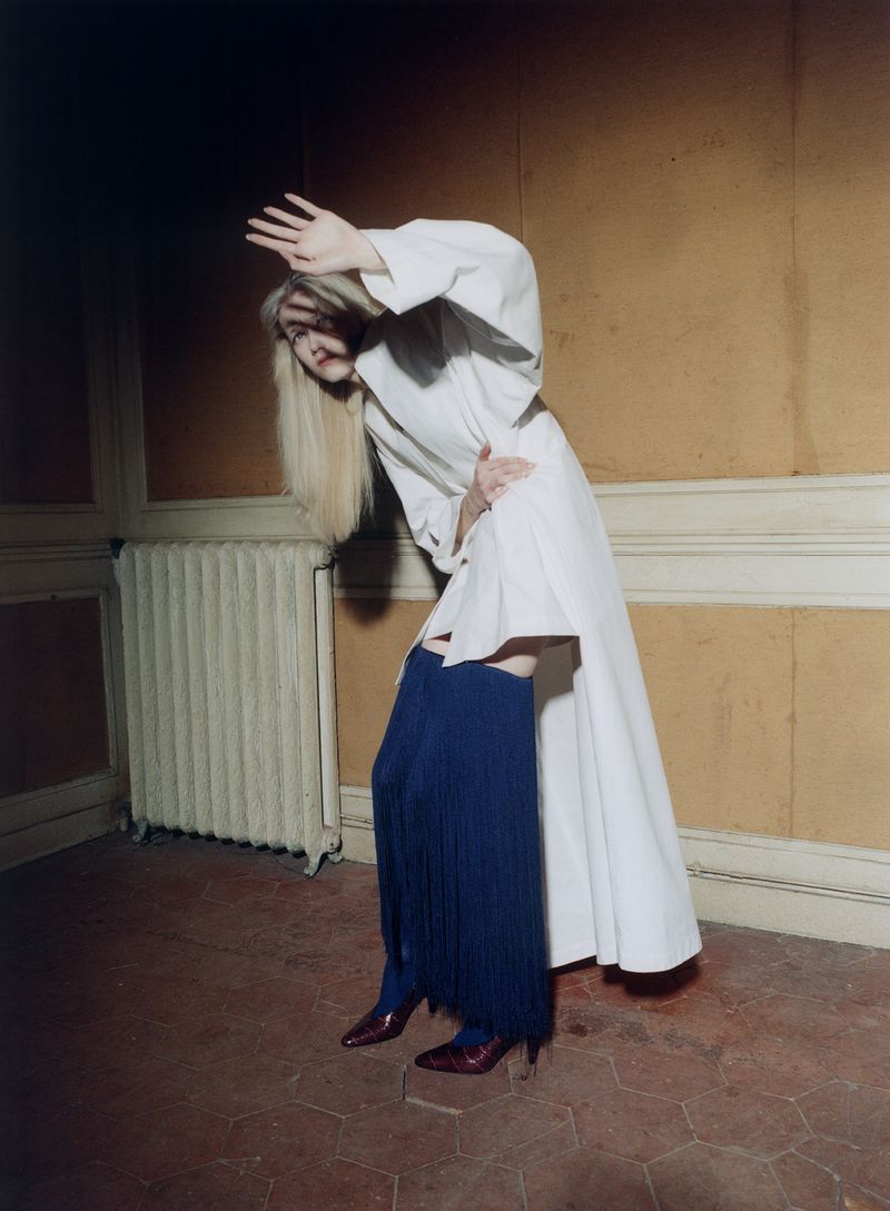 Model Malin Rudnick poses dynamically in a long white coat and blue fringe boots inside a room with ochre walls.