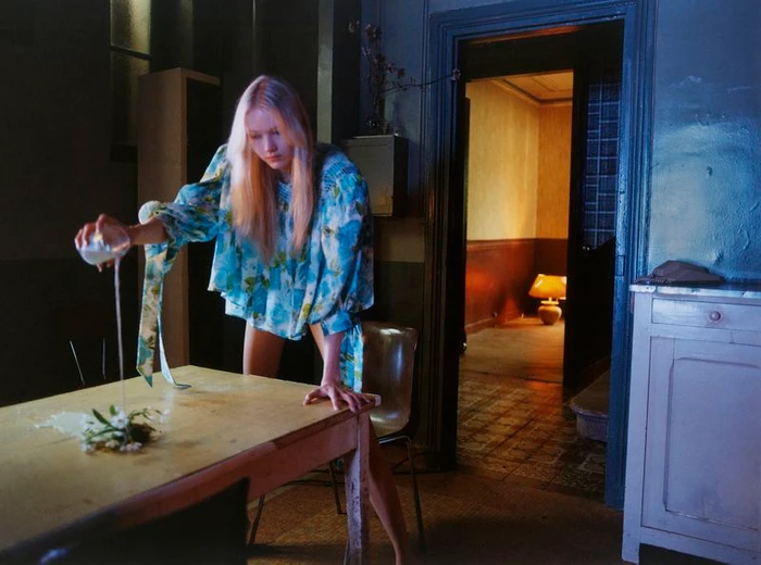 A blonde woman in a blue patterned top leans over a wooden table pouring white liquid onto flowers in a dimly lit room.