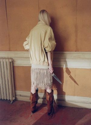 A woman seen from behind facing a wall, wearing a pale jacket, fringe skirt, and brown cowboy boots while holding a large knife.