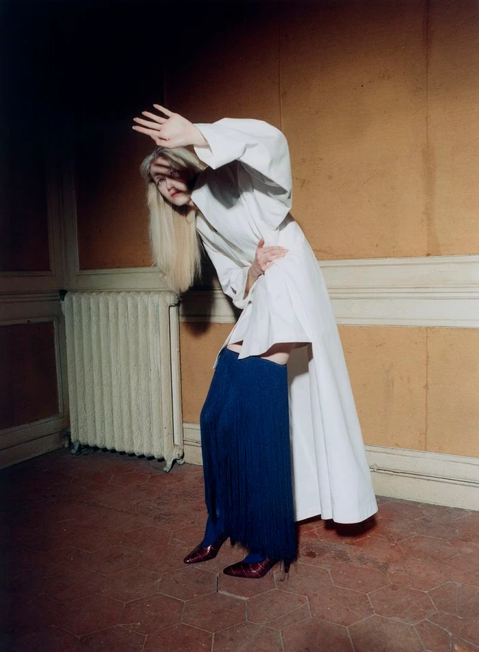 Model Malin Rudnick poses dynamically in a long white coat and blue fringe boots inside a room with ochre walls.